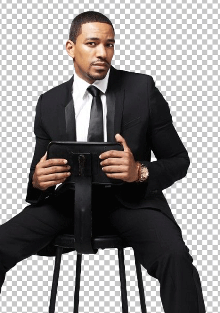 Laz Alonso is sitting on a stool, wearing a black suit and holding a tablet in his hand.