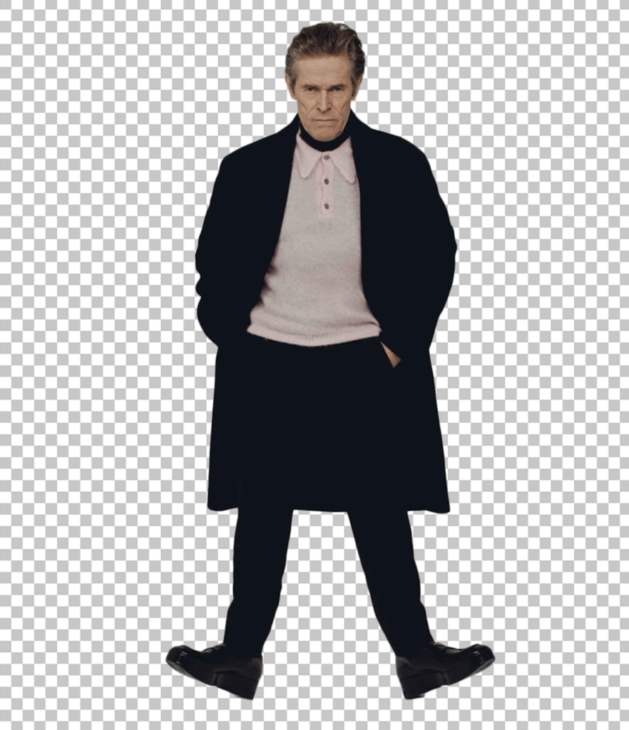 Willem Dafoe is wearing a black coat and pants and standing with his hands in his pocket.